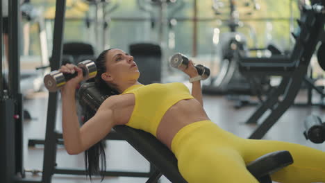 Determined-hispanic-female-athlete-doing-bench-press-with-dumbbells-in-gym.-A-fit-young-woman-doing-a-dumbbell-bench-press-in-a-small-gym.-Muscular-female-bodybuilder-lying-bench-and-doing-dumbbell